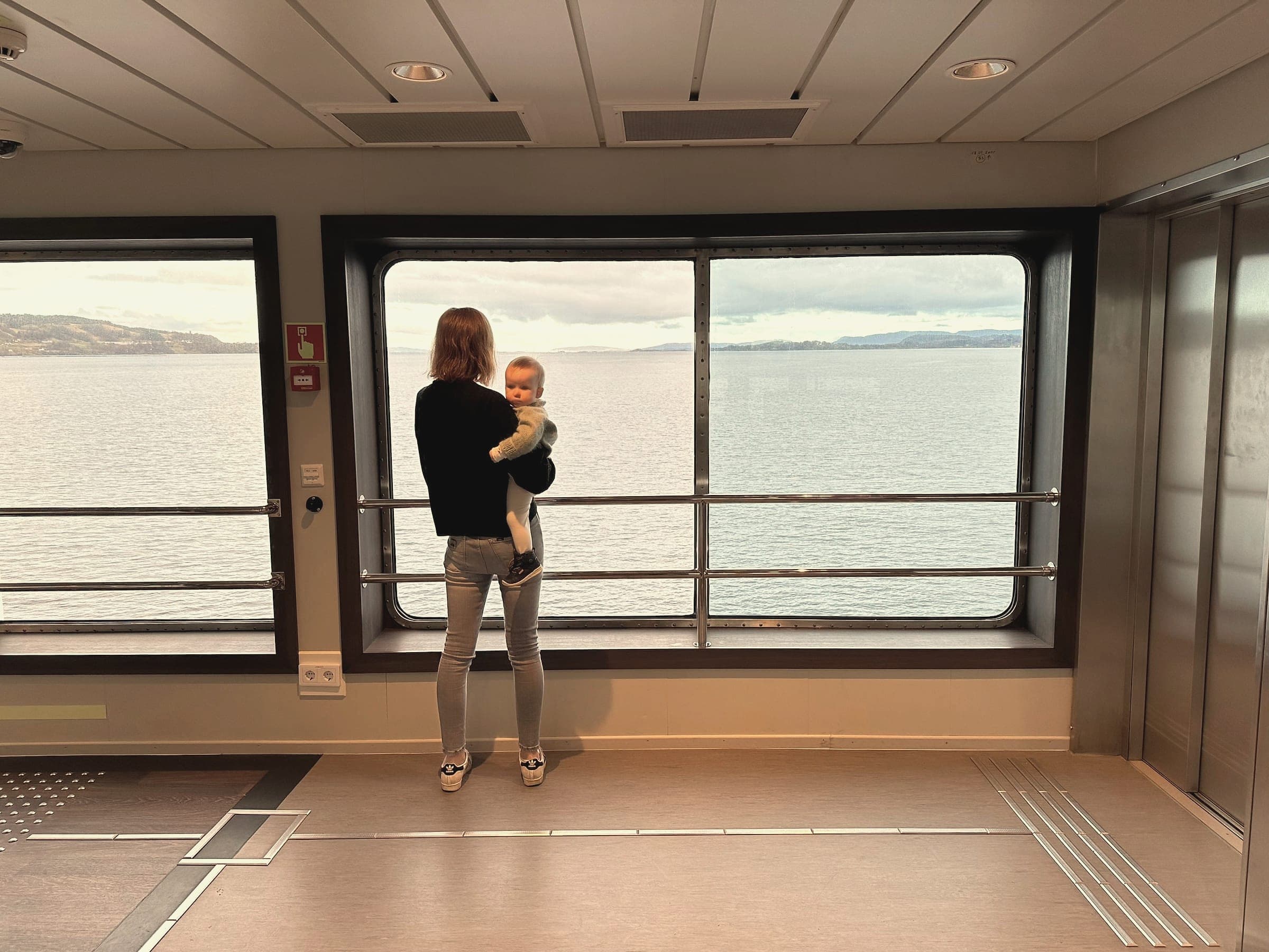 A parent holding a young child looks out a wide window toward calm water and distant hills.