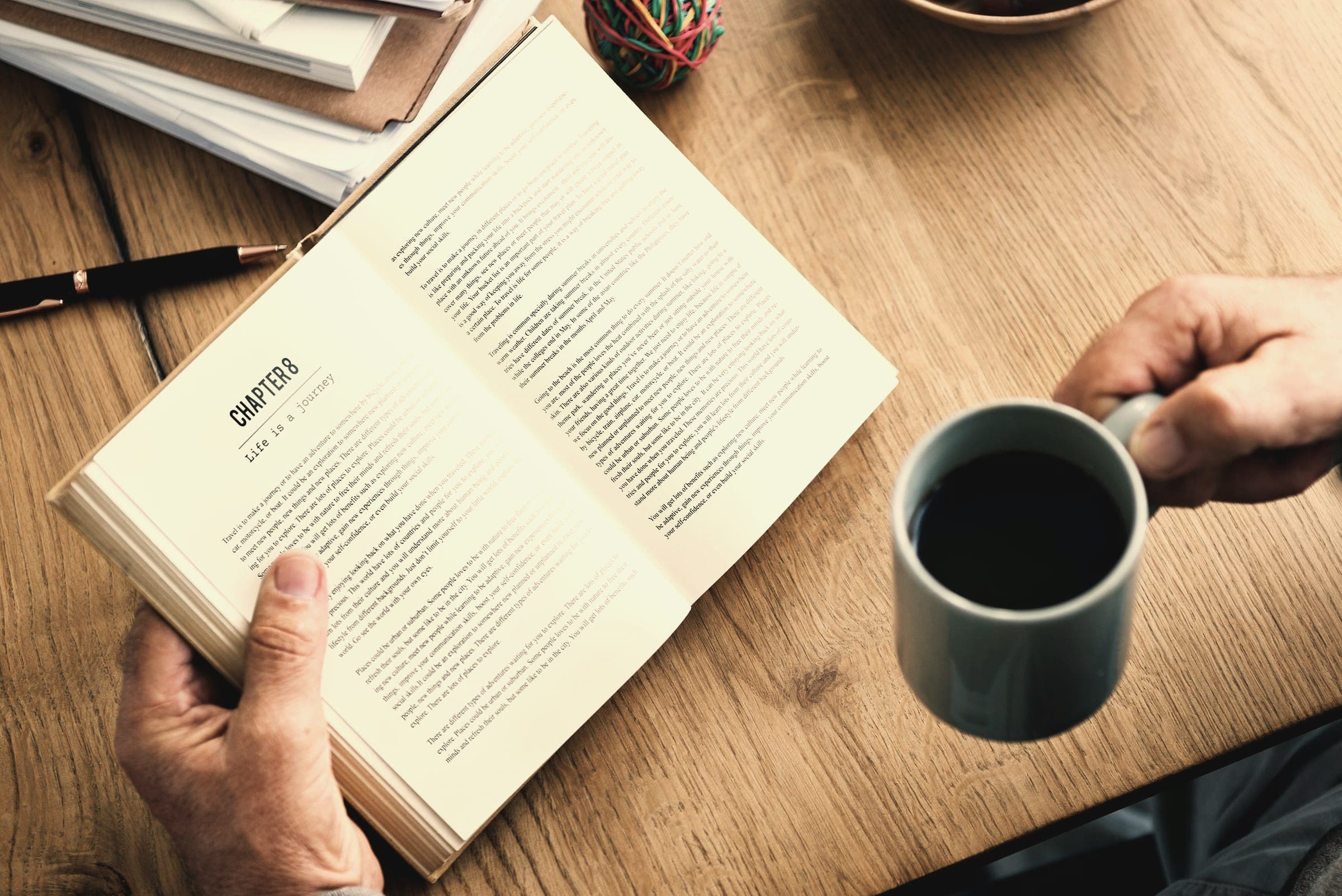 Hands hold an open book beside a cup of coffee on a wooden table.